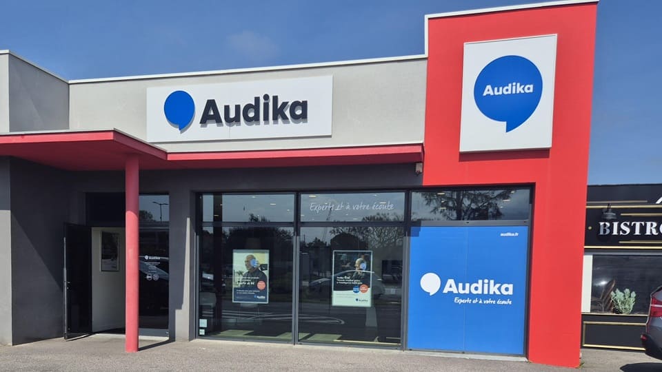 La devanture d’une clinique Audika, spécialisée dans les soins auditifs, affiche un logo bleu avec ’Audika’ et le slogan « Expert de votre écoute » sur une porte vitrée.