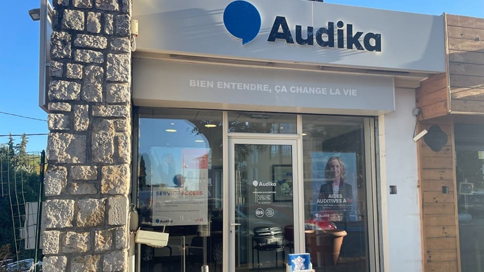 La façade de la clinique Audika présente une enseigne avec son logo bleu et noir, le slogan « BIEN ENTENDRE, ÇA CHANGE LA VIE ». Une porte vitrée et des affiches marketing, incluant « AIDES AUDITIVES À 0€ » et « SERVICE & ACCESS », sont visibles dans un cadre urbain.