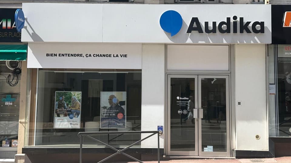 La façade d’une clinique Audika avec le slogan « BIEN ENTENDRE, ÇA CHANGE LA VIE », deux affiches promotionnelles de tests auditifs en vitrine, et une entrée avec porte vitrée.