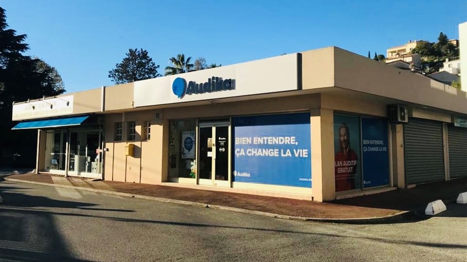 La façade d'une clinique Audika, spécialisée dans les soins auditifs, affiche le logo bleu 'Audika' et les slogans : 'BIEN ENTENDRE, ÇA CHANGE LA VIE' et 'UN AUDIT GRATUIT.' Contextualisée dans un environnement urbain ensoleillé.