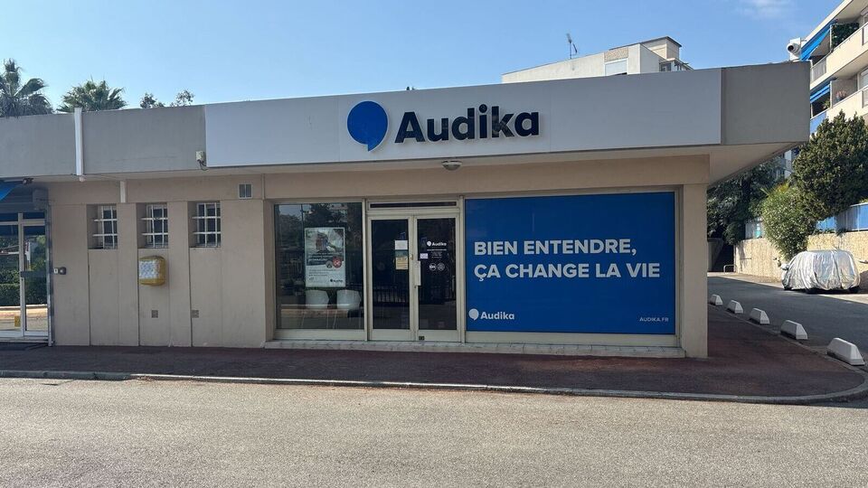 La façade d'une clinique Audika, spécialisée dans les soins auditifs, affiche le logo bleu 'Audika' et les slogans : 'BIEN ENTENDRE, ÇA CHANGE LA VIE' et 'UN AUDIT GRATUIT.' Contextualisée dans un environnement urbain ensoleillé.