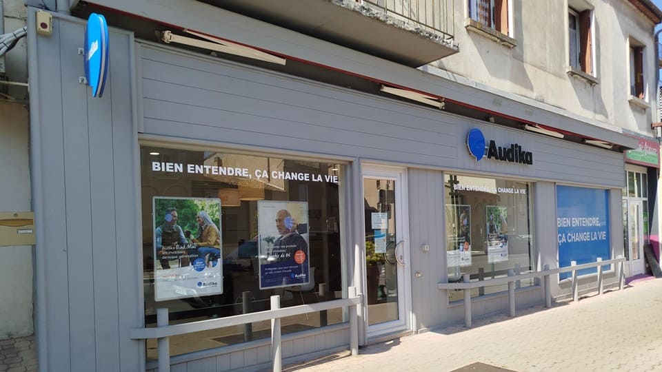 La façade d’une clinique Audika grise, spécialisée en soins auditifs, affiche le slogan « Bien entendre, ça change la vie ». Des affiches promeuvent des appareils auditifs et services.