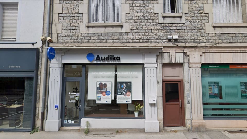 La façade d’un centre Audika arbore un logo bleu et noir, avec une vitrine affichant des posters promotionnels. Texte visible : « BIEN ENTENDRE, ÇA CHANGE LA VIE ». Environnement urbain.