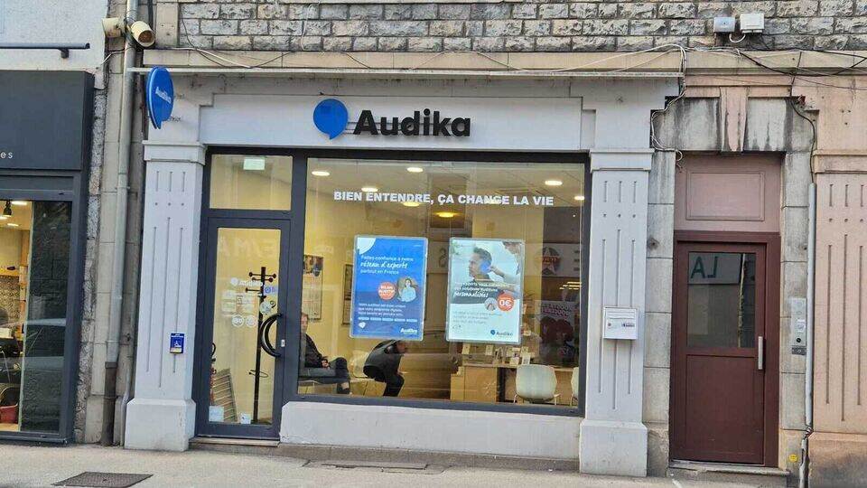 La façade d’un centre Audika arbore un logo bleu et noir, avec une vitrine affichant des posters promotionnels. Texte visible : « BIEN ENTENDRE, ÇA CHANGE LA VIE ». Environnement urbain.