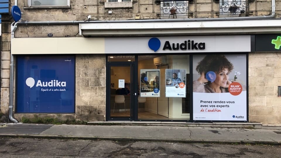 La façade d’un centre Audika montre une vitrine avec le logo « Audika, Experts à votre écoute » sur fond bleu, des affiches promotionnelles sur l’audition, et l’inscription : « Prenez rendez-vous avec vos experts de l’audition - Bilan auditif gratuit ». L’enseigne se situe dans un bâtiment en pierre, avec une porte vitrée centrale et des sièges visibles à l’intérieur, suggérant un accueil chaleureux pour des tests auditifs et conseils spécialisés.