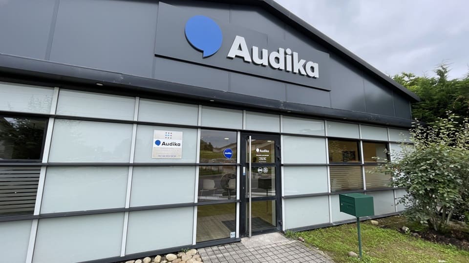 Un bâtiment moderne abritant une clinique Audika, spécialisé en soins auditifs. La façade vitrée montre le logo d’Audika, avec un panneau indiquant ’Audika - Votre meilleur partenaire auditif’.