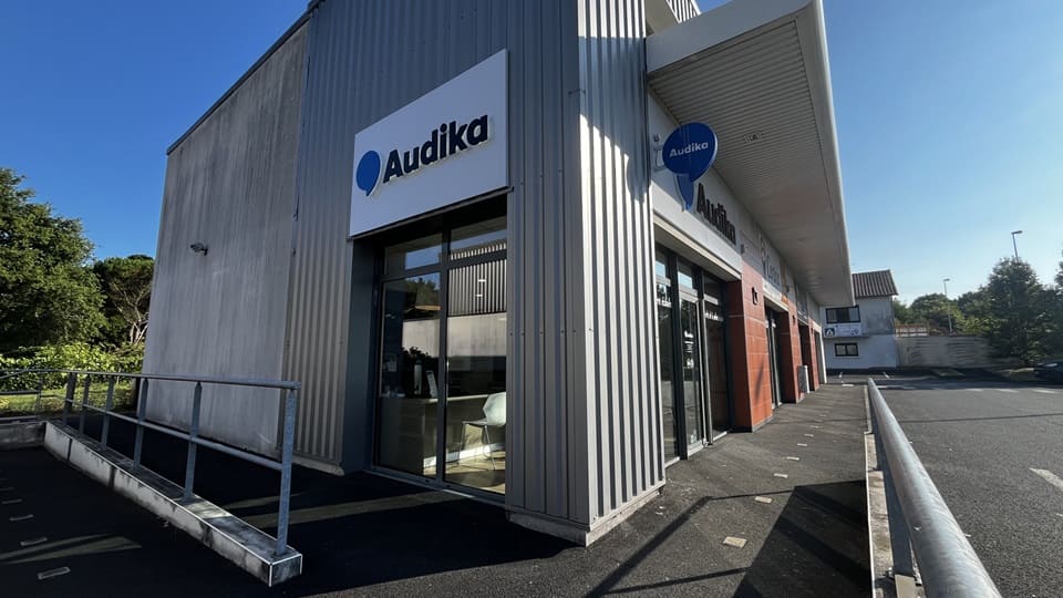 Façade d'une clinique Audika spécialisée en soins auditifs, présentant une entrée vitrée avec un logo clairement visible. Environnement calme, parking adjacent et rampe d'accès accessible.