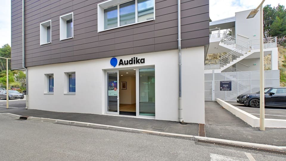 La façade d’une clinique Audika, spécialisée dans les soins auditifs, avec une entrée vitrée et l’inscription ’Audika’. Contexte : un bâtiment moderne avec parking et escaliers extérieurs.