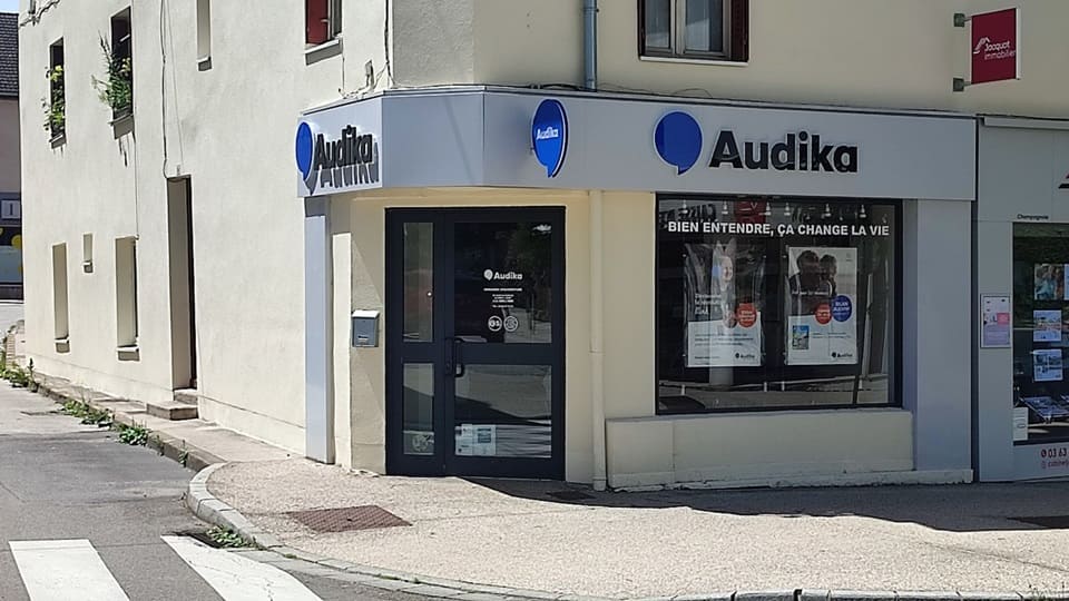 Une façade de centre auditif Audika avec logo bleu et texte 'Bien entendre, ça change la vie,' située dans une rue ensoleillée, affichant des posters promotionnels derrière une vitrine.