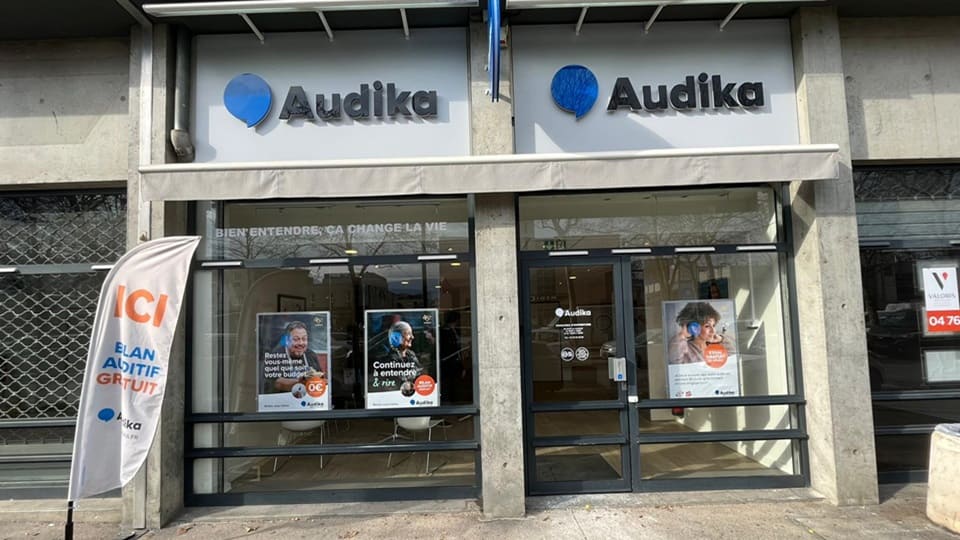 La façade d'une clinique Audika spécialisée en soins auditifs montre de grandes vitrines avec affiches promotionnelles. Texte visible : « Bien entendre, ça change la vie » et « ICI – BILAN AUDITIF GRATUIT ».