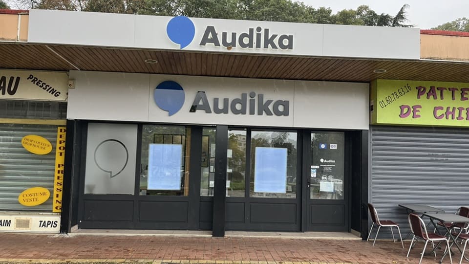 La façade d'une clinique Audika, spécialisée en soins auditifs, présente une enseigne blanche avec logo bleu. Contexte : environnée de commerces, avec terrasse de café à droite.