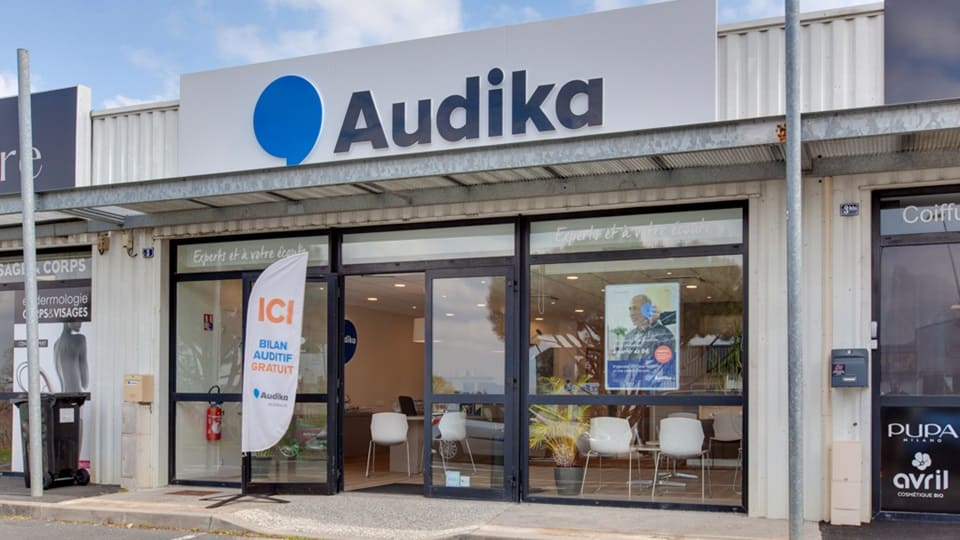 La façade d’un centre Audika, spécialisé en soins auditifs, affiche un panneau 'ICI! Bilan auditif gratuit'. L’intérieur moderne est visible, avec des chaises blanches. Environnement commercial urbain.