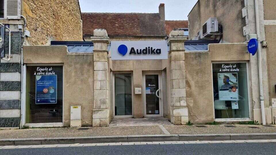 La façade d’un centre auditif Audika avec vitrine affichant ’Experts et à votre écoute.’ Contenu promotionnel en vitrine, porte centrale, murs en pierre dans une rue calme.