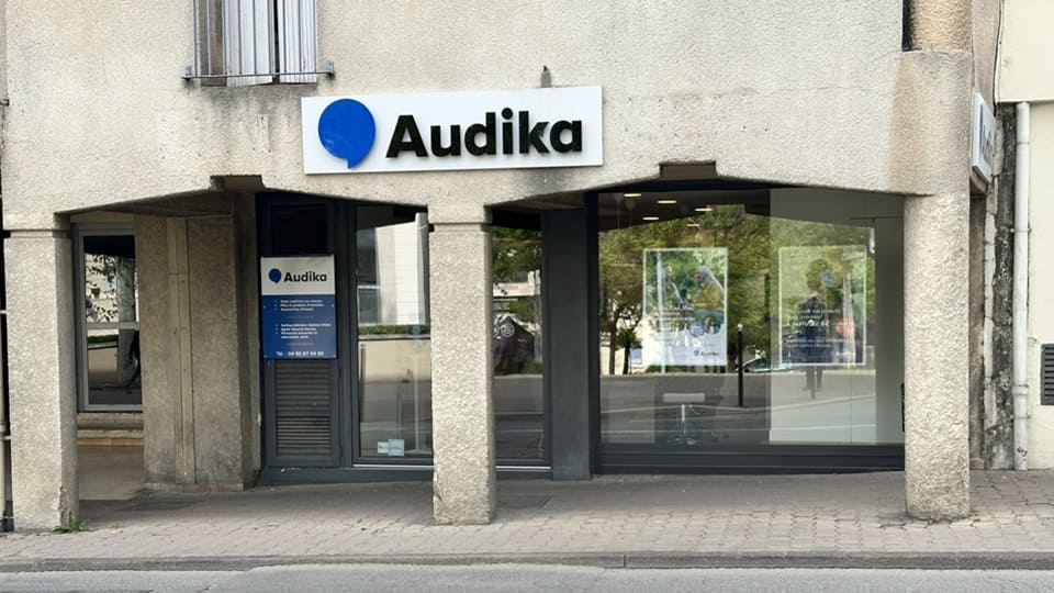 La façade d’une clinique Audika montre une enseigne bleue et blanche avec le logo. Deux affiches publicitaires visibles à travers les vitrines promeuvent des soins auditifs.