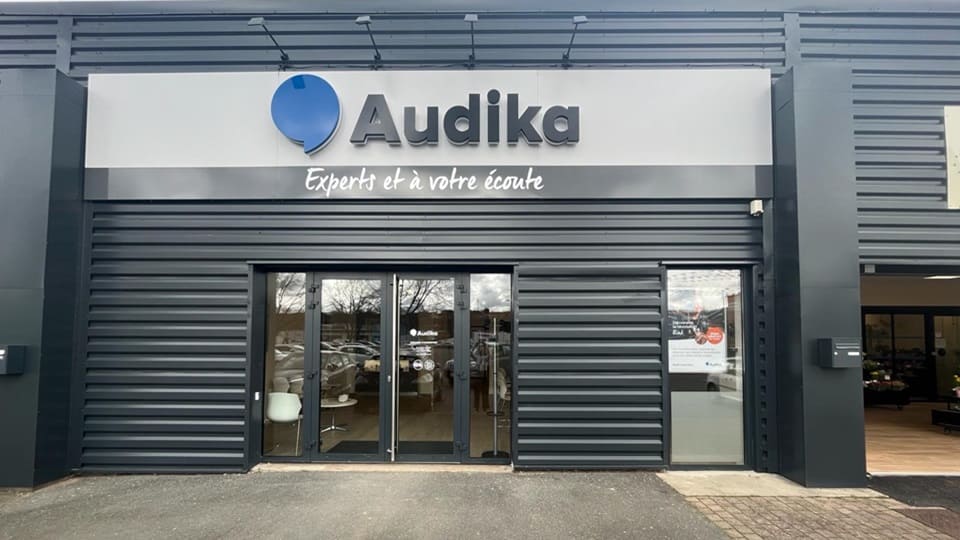 Une façade moderne de clinique Audika, avec le logo bleu et texte « Experts et à votre écoute ». Contexte : vitrines transparents montrant sièges, parking extérieur reflété.