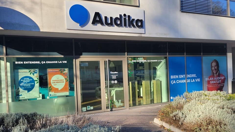La façade d'une clinique Audika, spécialisée en soins auditifs, affiche le slogan 'Bien entendre, ça change la vie' et propose un 'Bilan auditif gratuit'. Des affiches colorées promeuvent parrainages et bilans auditifs.