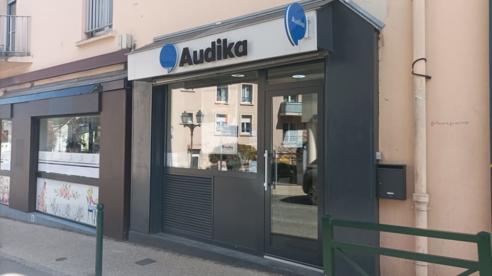 La façade d’un centre auditif Audika avec enseigne blanche et logo bleu, des vitrines modernes et une porte vitrée, située dans un quartier urbain aux façades claires.
