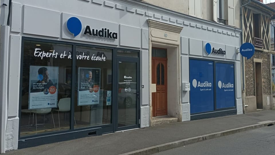 La devanture d’une clinique Audika affiche le logo bleu, le slogan « Experts et à votre écoute », des affiches promotionnelles, et des vitrines bleues dans une rue résidentielle.
