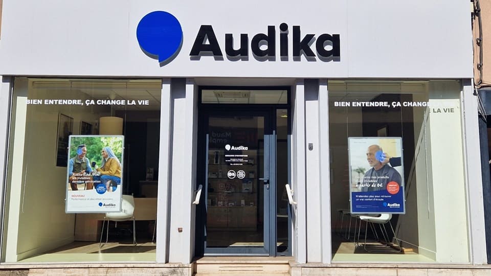 La façade d’une clinique Audika présente un logo bleu et noir, avec la phrase ’BIEN ENTENDRE, ÇA CHANGE LA VIE’. Deux affiches publicitaires montrent des services pour aides auditives.
