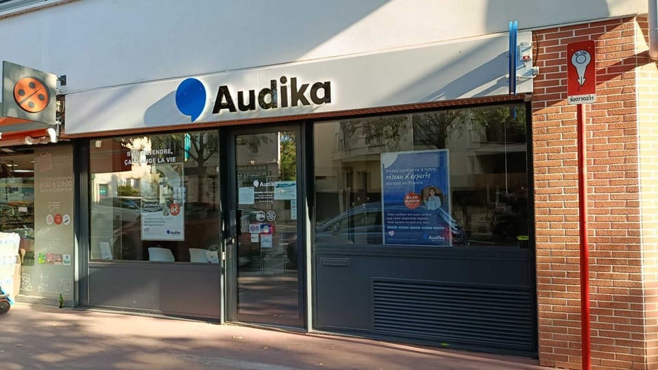 La façade d'une clinique Audika spécialisée dans les soins auditifs, affiche le texte : « BIEN ENTENDRE, ÇA CHANGE LA VIE » et « AIDES AUDITIVES À 0€' ». Une affiche met en avant des prothèses auditives signant la présence d'audioprothésistes. Le cadre urbain inclut du mobilier moderne et une vitrine lumineuse.
