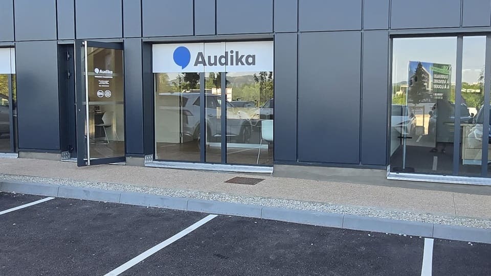 La façade d’une clinique Audika spécialisée en soins auditifs présente des vitrines transparentes, une porte ouverte, un logo bleu et blanc ’Audika,’ et un parking adjacent. Aucun texte supplémentaire visible.