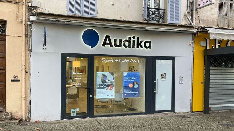 La devanture d’une clinique Audika affiche le logo bleu et noir de l’entreprise, avec des affiches promotionnelles sur les aides auditives et services d’audiologie visibles à travers les vitrines.
