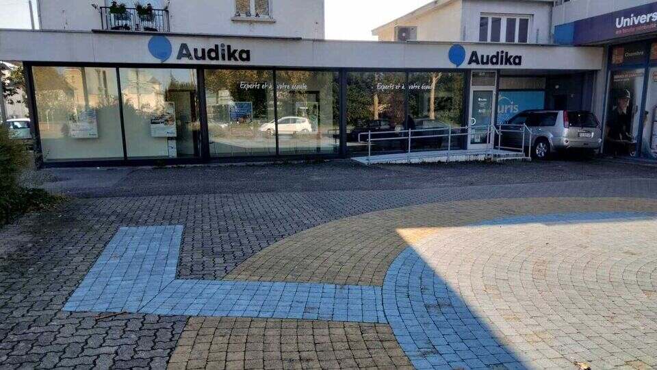 La façade d’une clinique Audika affiche le logo bleu de l’enseigne. Des affiches publicitaires sur les appareils auditifs décorent les grandes vitrines. Le bâtiment est situé dans un quartier résidentiel.