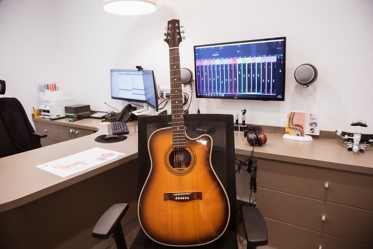 Guitare acoustique posée sur un fauteuil dans un cabinet d’audioprothésiste, avec casque audio et équipements professionnels en arrière-plan.