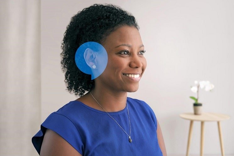 Une femme portant une robe bleue est photographiée dans un environnement lumineux et minimaliste. Une image en surimpression souligne son oreille, suggérant un lien avec un appareil auditif. Un tabouret avec une plante en pot est visible en arrière-plan. Aucun texte n’apparaît dans l’image.