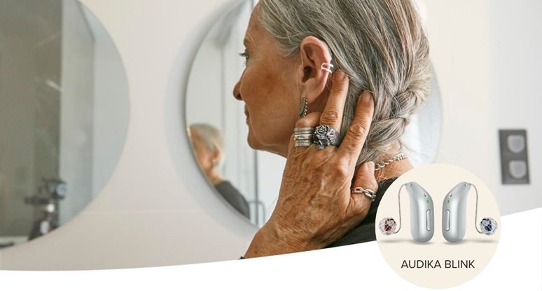 Femme équipée d’aides auditives Audika Blink ajustant ses cheveux devant un miroir.