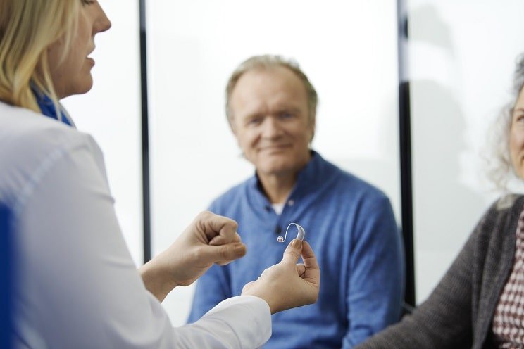 Un audioprothésiste explique le fonctionnement d’un appareil auditif à un couple lors d’une consultation.