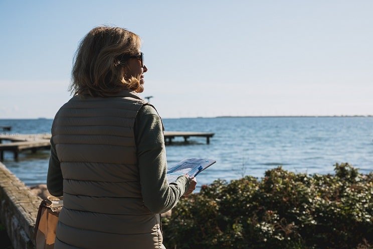 Femme de dos tenant un magazine près de la mer, avec un quai et un horizon dégagé en arrière-plan.