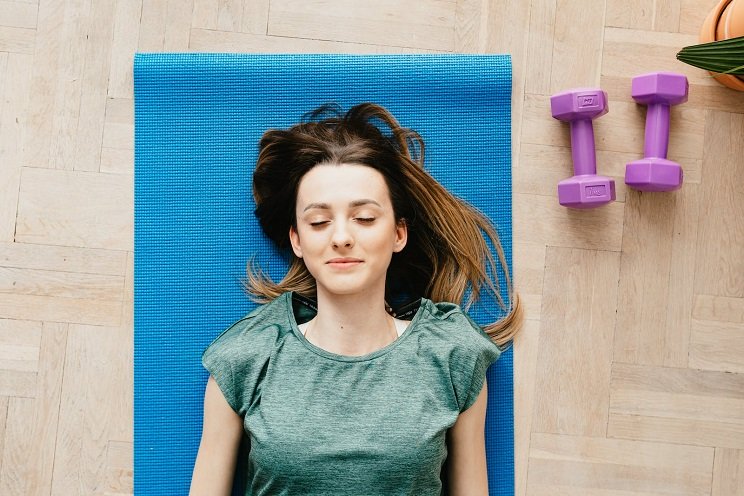 Une femme allongée sur un tapis de yoga bleu, portant un t-shirt vert, se repose après une activité physique; à droite, deux haltères violets sont posés sur un sol en bois.
