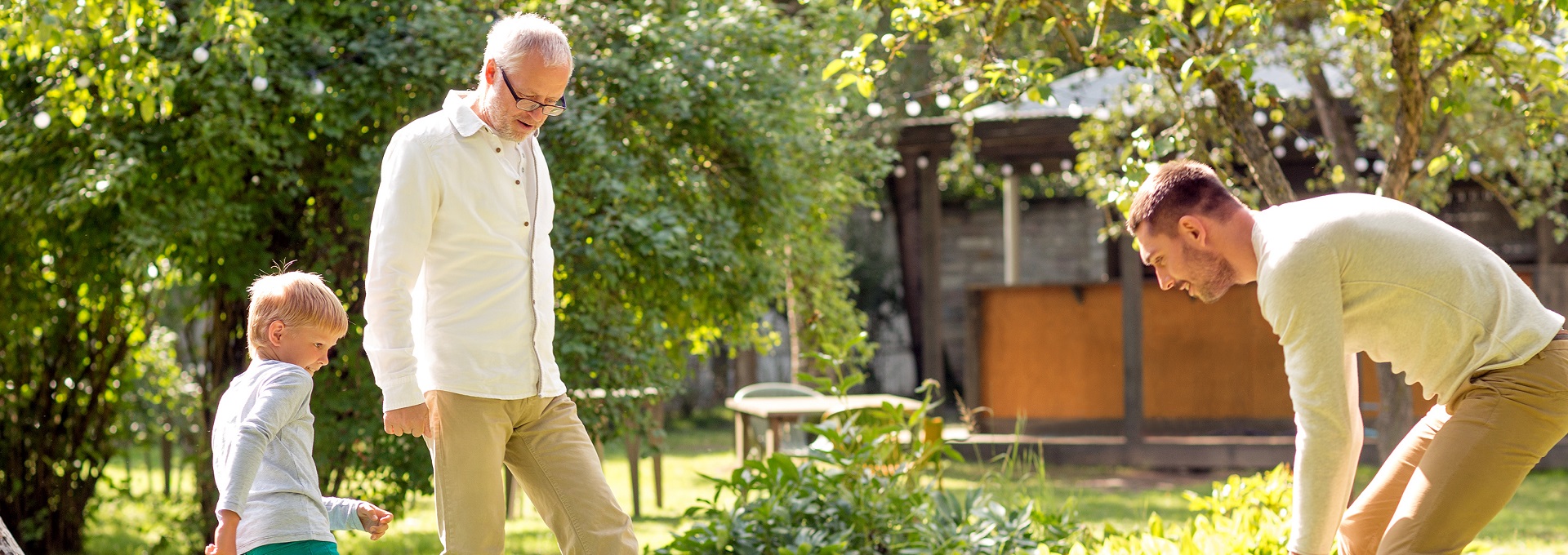 Un homme âgé, un jeune homme et un enfant jouent ensemble dans un jardin ensoleillé entouré de plantes, d’arbres et de structures en bois, créant une atmosphère familiale conviviale.