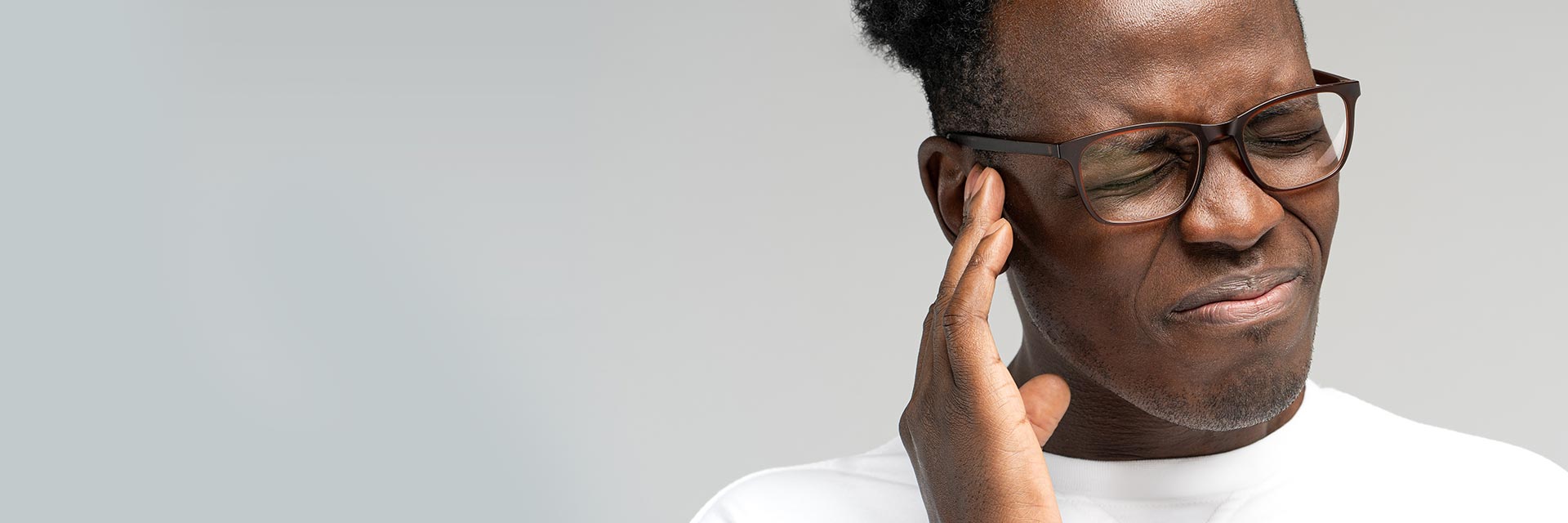 Une main touche l'oreille d'une personne portant des lunettes. Contexte neutre avec un fond gris clair. Aucune mention explicite d'appareils auditifs ou de texte visible.
