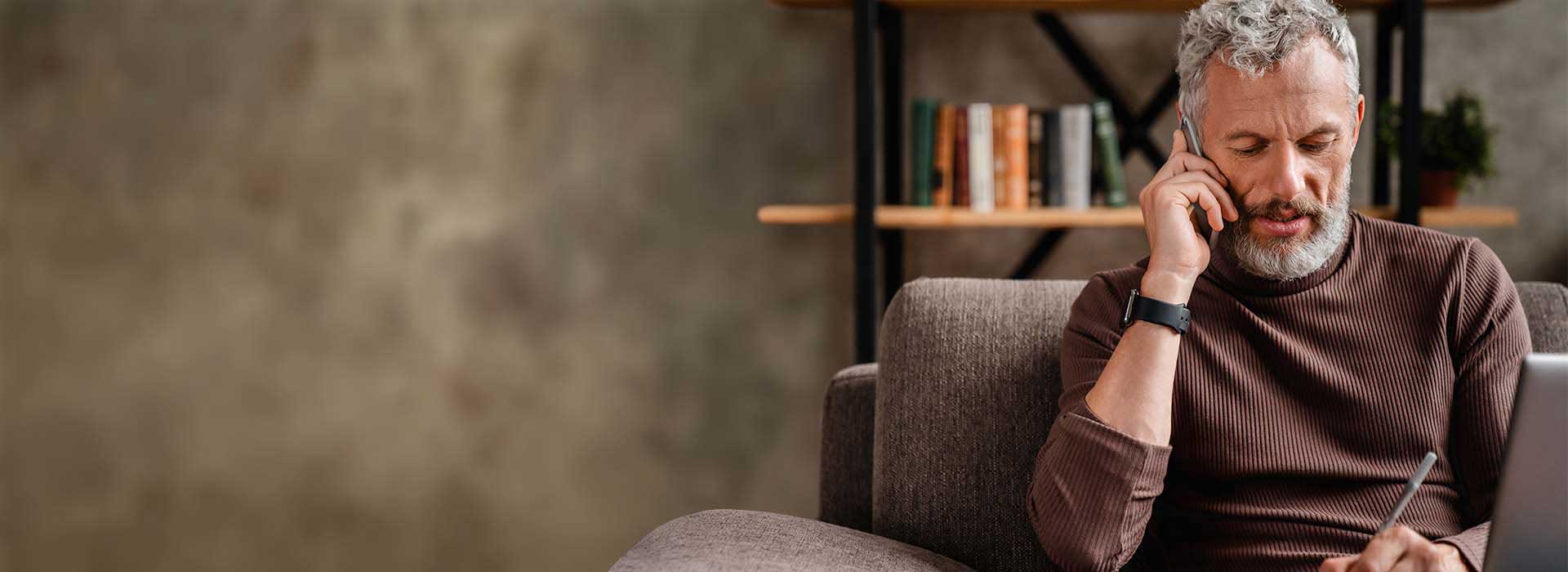 Un homme portant un appareil auditif (marque non identifiable) touche son oreille tout en tenant un stylo. Il est assis sur un canapé dans un intérieur chaleureux avec une bibliothèque en arrière-plan.