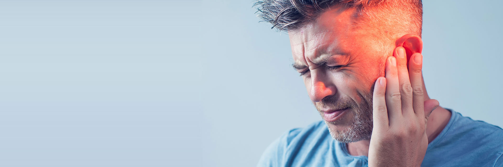 Un homme touche son oreille droite, visiblement douloureuse avec une lumière rouge accentuant l’inconfort, dans un contexte sobre sur fond bleu clair, évoquant des problèmes auditifs ou une consultation auditive.