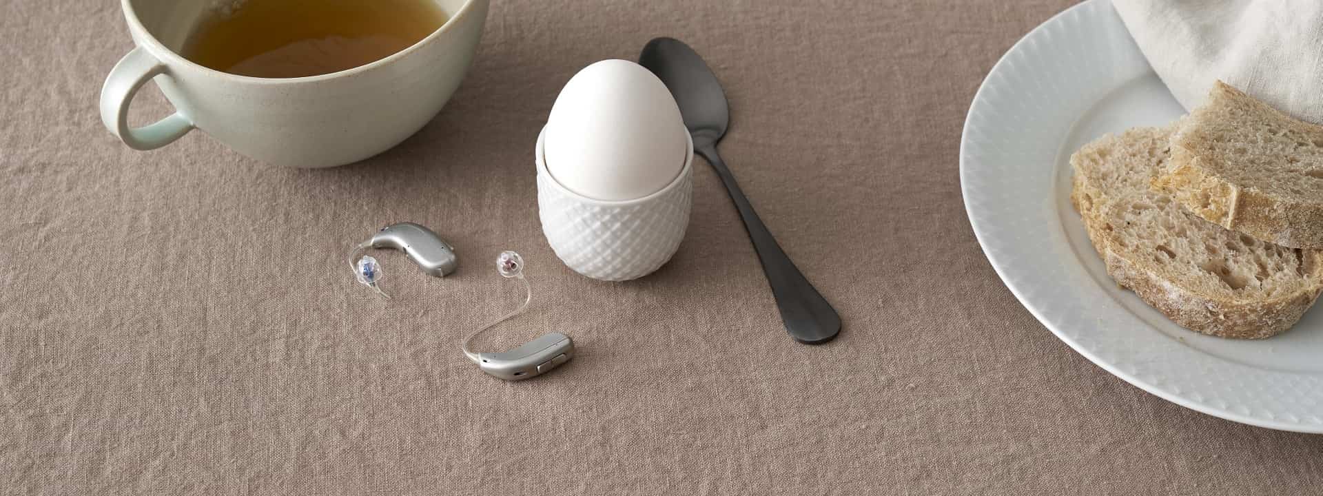 Appareils auditifs avec embouts colorés posés sur une table près d’un œuf à la coque, une cuillère, une tasse de thé et des tranches de pain.