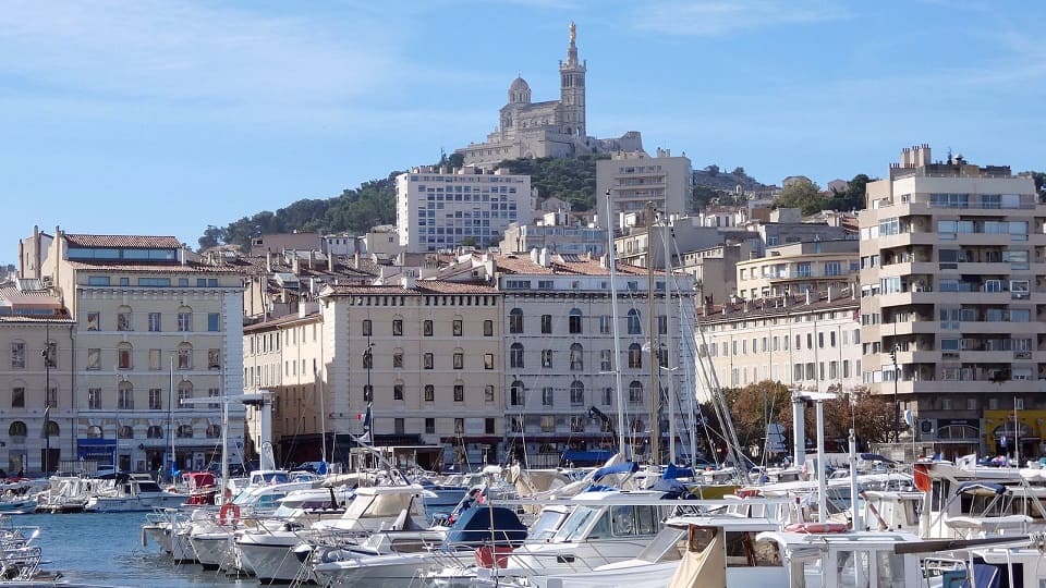 Vue sur le Vieux-Port de Marseille avec des bateaux au premier plan et Notre-Dame-de-la-Garde au sommet de la colline en arrière-plan.