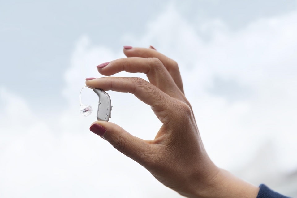 Appareil auditif moderne tenu entre deux doigts devant un ciel dégagé.