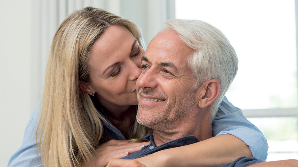 Une femme blonde embrasse tendrement la joue d’un homme aux cheveux gris, tous deux souriants et enlacés dans un moment chaleureux.