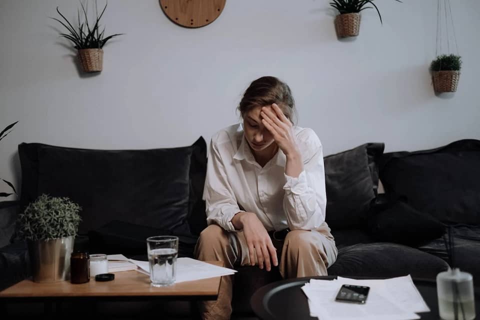 Une jeune femme en chemise blanche assise sur un canapé, tenant sa tête avec une expression de malaise, entourée de documents et de verres sur une table basse, symbole d'un inconfort lié aux acouphènes.