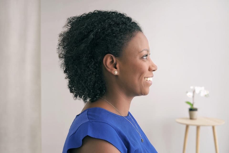 Une femme de profil portant un haut bleu et des boucles d’oreilles perle; en arrière-plan, une table en bois avec une orchidée blanche dans un pot minimaliste, devant un mur clair.