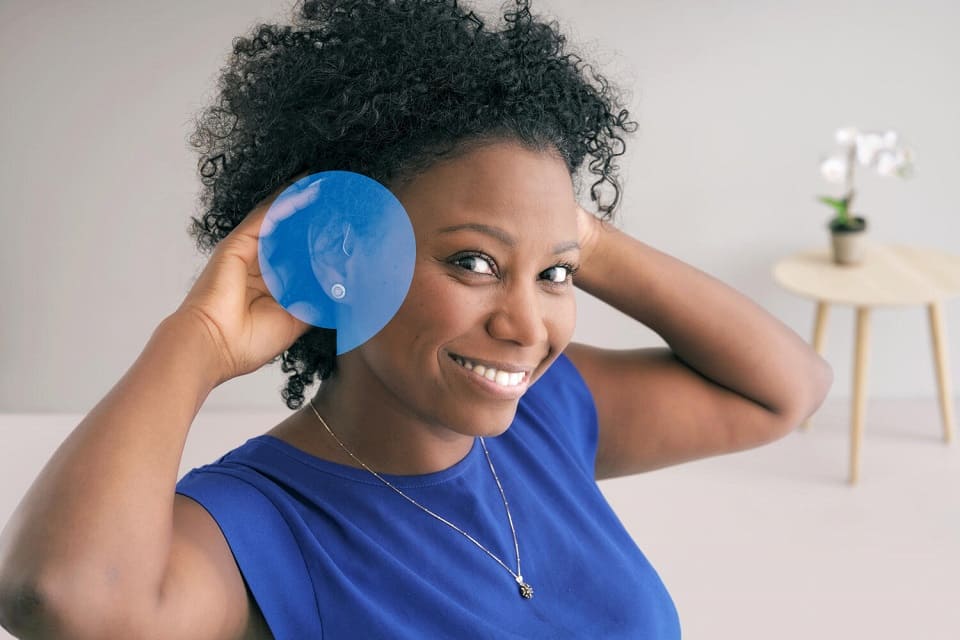Une personne tenant un objet bleu contre son oreille gauche, portant une robe bleue, dans un environnement lumineux avec une table en bois et une orchidée blanche en arrière-plan.