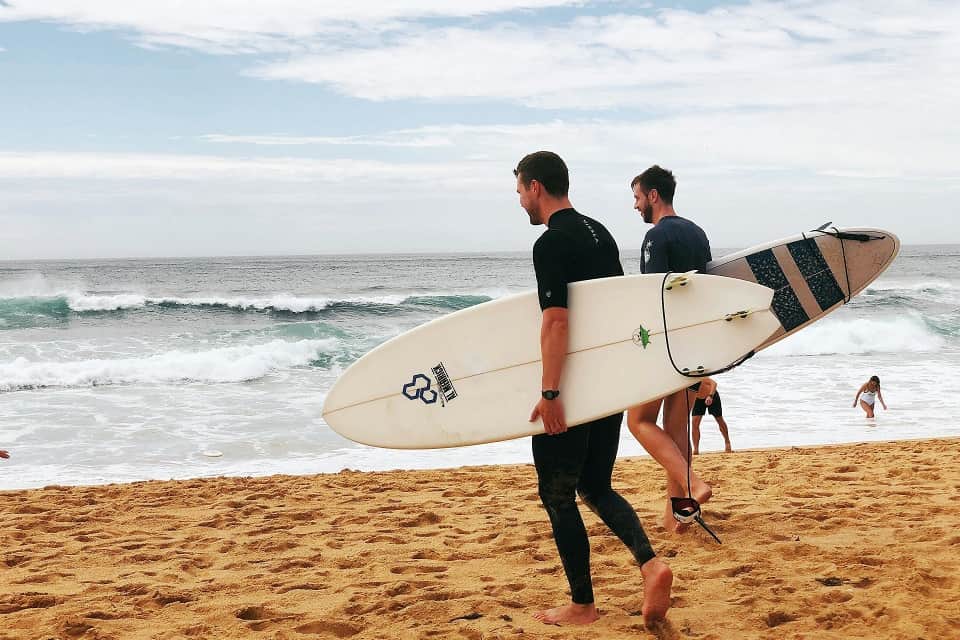 Deux surfeurs marchent sur la plage avec leurs planches de surf, l’océan en arrière-plan.