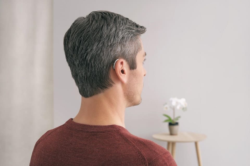 Un homme aux cheveux poivre et sel porte un appareil auditif discret derrière l’oreille gauche. Il est assis dans une pièce lumineuse avec une petite table et une orchidée blanche.