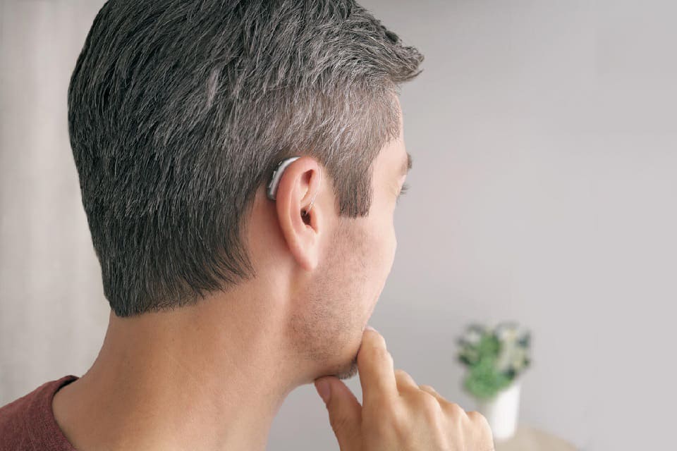 Un homme porte une prothèse auditive discrète derrière l'oreille gauche, probablement d'Oticon, Bernafon ou Philips, dans un intérieur lumineux avec une plante en pot visible en arrière-plan.