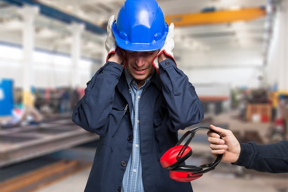 Un ouvrier portant un casque bleu se couvre les oreilles dans un atelier bruyant, tandis qu'une main lui tend un casque antibruit rouge.
