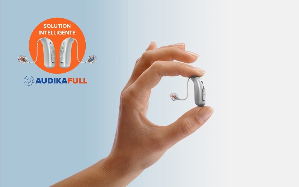 Une main tient un appareil auditif moderne avec le logo Audika Full et le texte ’Solution intelligente’ en arrière-plan.