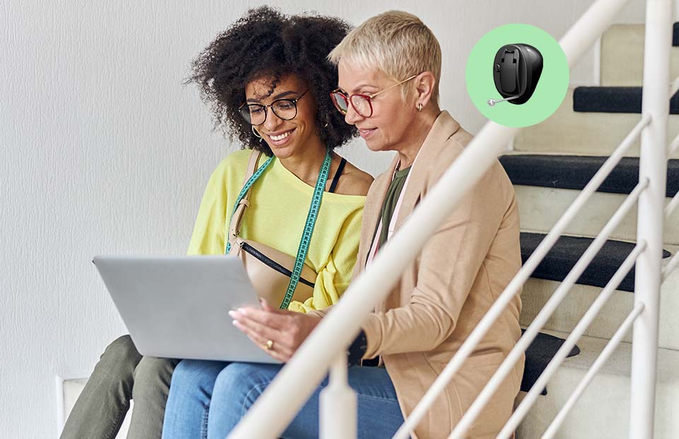 Deux femmes assises sur un escalier discutent tout en regardant un ordinateur portable, avec un appareil auditif numérique affiché à côté.